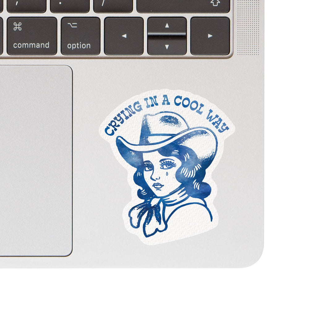 Laptop with a sticker of a woman in a cowboy hat and text 'Crying in a Cool Way' on a white background