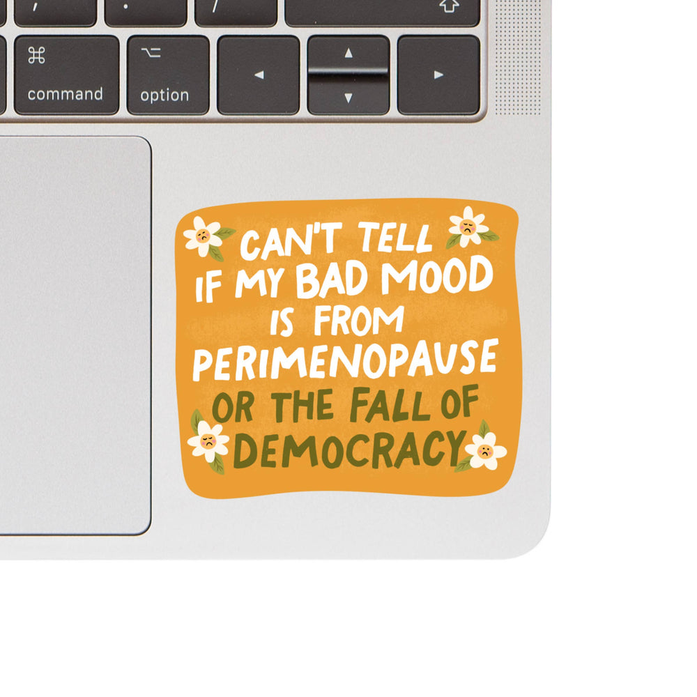 Laptop with a sticker reading 'Can't tell if my bad mood is from perimenopause or the fall of democracy' on a white background.