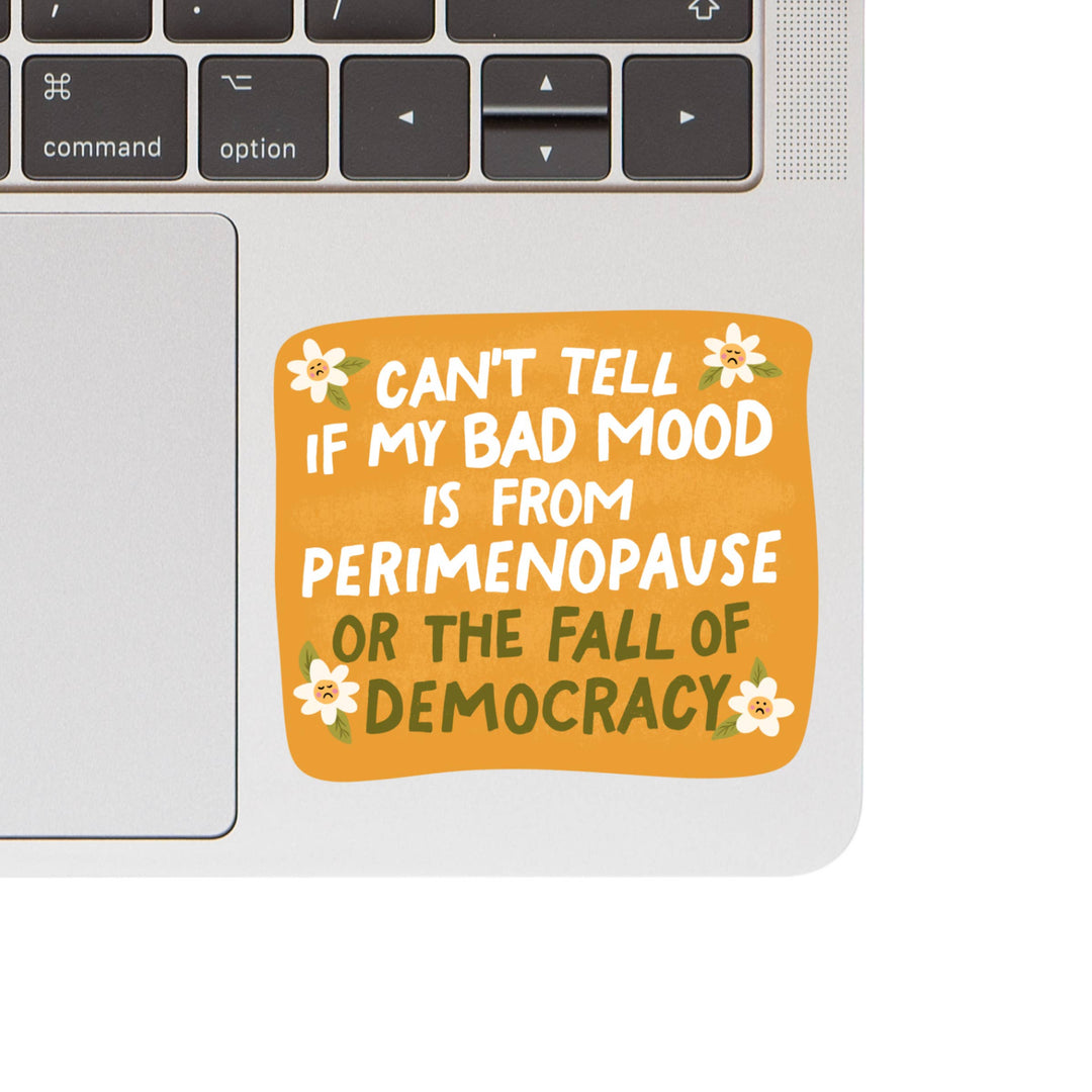Laptop with a sticker reading 'Can't tell if my bad mood is from perimenopause or the fall of democracy' on a white background.